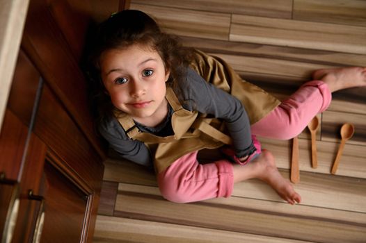 Top view pretty little girl, cute daughter in pink pants, gray long sleeve, beige chef apron and kitchen mittens, sitting barefoot on floor with scattered wooden spoons, looking dreamily up at camera