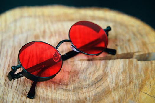 Round sunglasses with red lens on a wooden stand. Fashionable unisex party accessory. Hippie or boho style.