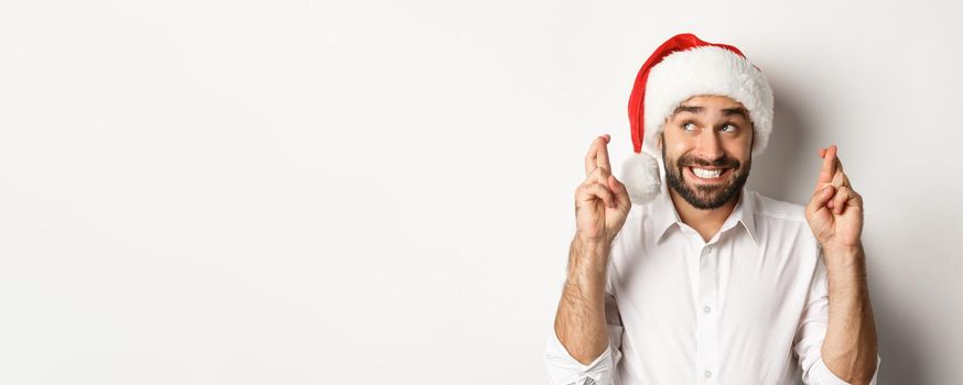 Party, winter holidays and celebration concept. Happy man in santa hat making christmas wish, cross fingers for good luck and smiling excited, white background.