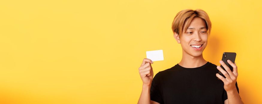 Close-up of handsome stylish asian guy shopping online, looking at mobile phone and smiling, showing credit card, standing over yellow background.