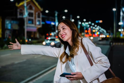 Beautiful woman smiling using smartphone application hailing with hand up calling cab after late work, Asian businesswoman standing hail waving hand taxi on road in busy city street at night