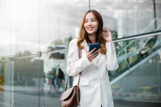 Portrait successful business woman smiling holding smartphone use application chat online in morning, Asian businesswoman working with mobile phone standing against street front building near office