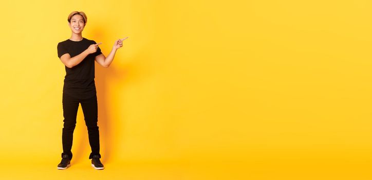 Full-length of handsome confident asian guy smiling happy, pointing fingers right, showing logo, yellow background.