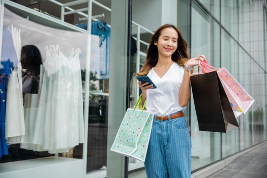 Smiling shopper young girl lifestyle standing using mobile phone after shopping on city street, Happy Asian beautiful woman holding shopping bag and smartphone on her hands near the mall shop window