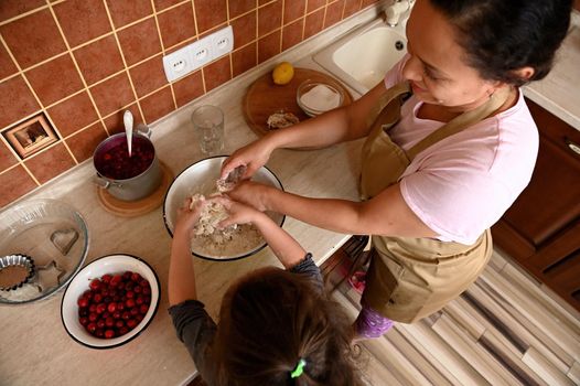 Overhead view of a dark-haired multi-ethnic beautiful woman, a loving caring mother and her adorable little daughter, enjoying cooking together in the home kitchen, kneading dough for a cherry pie