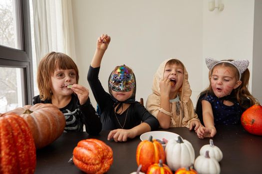 Long haired junior schoolgirls and preschooler siblings in Halloween costumes eat treats sitting at table decorated with pumpkins at home