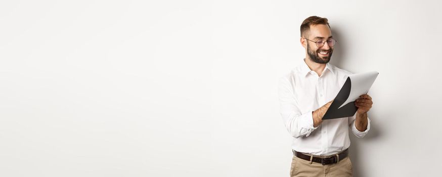 Man looking satisfied while reading documents, holding clipboard and smiling, standing over white background.