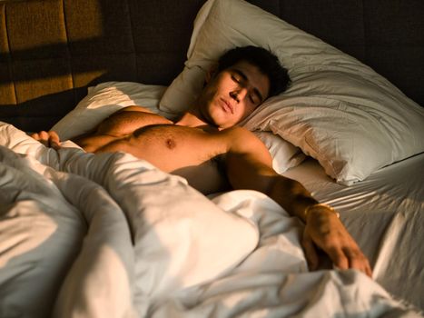 Handsome shirtless athletic young man laying in bed at night with eyes closed, sleeping