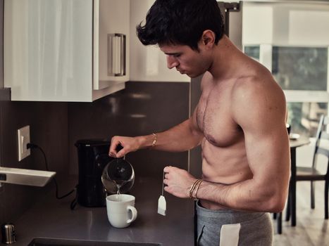 Side view of young shirtless male with tea bag pouring water in mug while brewing drink for breakfast in morning at home