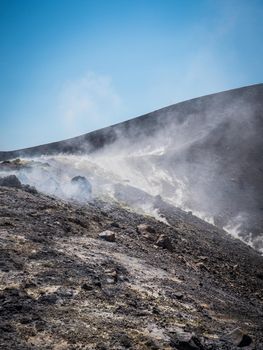 Hilly uneven slopes with rough boulders and smoke, on volcanic island Vulcano, located in Southern Italy in sunny day with blue sky