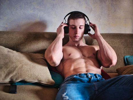 Handsome shirtless young muscular bodybuilder man listening to music with headphones while sitting on couch