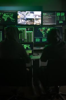 Two hooded hackers hacking security firewall late at night in basement hideout. Vertical image. Surveillance team.