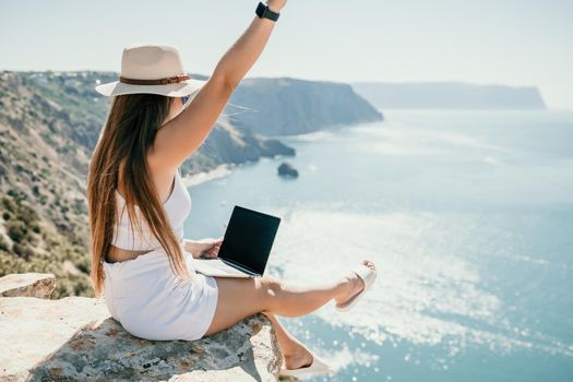 Successful business woman in yellow hat working on laptop by the sea. Pretty lady typing on computer at summer day outdoors. Freelance, travel and holidays concept.