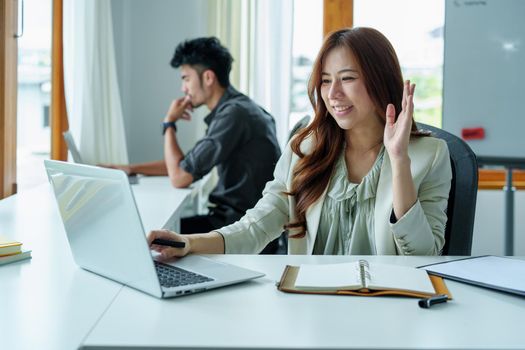 Portrait of an Asian bank employee using a laptop video conferencing with team members working on budget paperwork at their desk in their office.