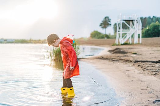 Boy in a red raincoat and yellow rubber boots playing with water at the beach. School kid in a waterproof coat jumping in water at sea. Child having fun with waves at the shore.