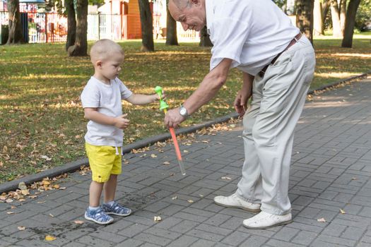 An old grandfather plays with his grandson on the weekend in an amusement park and cheerfully blow soap bubbles. An elderly man, a pensioner and a little boy spend time together.