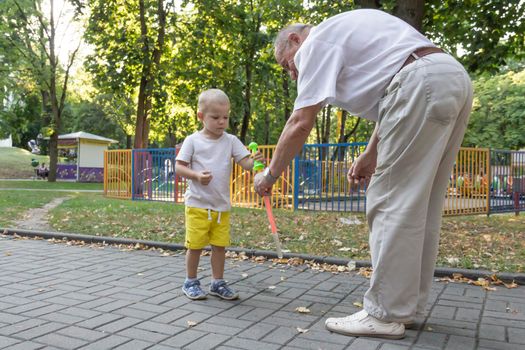 An old grandfather plays with his grandson on the weekend in an amusement park and cheerfully blow soap bubbles. An elderly man, a pensioner and a little boy spend time together.