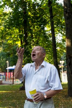 An elderly man walks alone in the park in the summer. The pensioner cheerfully throws popcorn up from a glass and catches it with his mouth wide open.