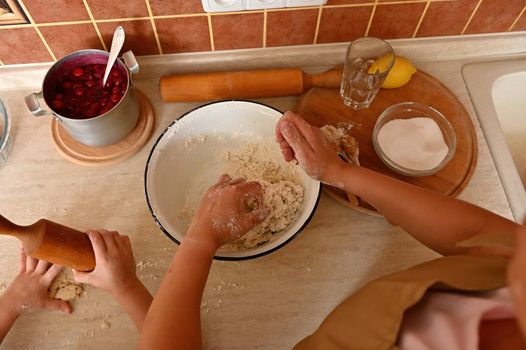 Top view of kitchen countertop with a saucepan of caramelized cherries for filling the pie and tartlets, hands of mom and child knead and roll out the dough with a wooden rolling pin. Selective focus