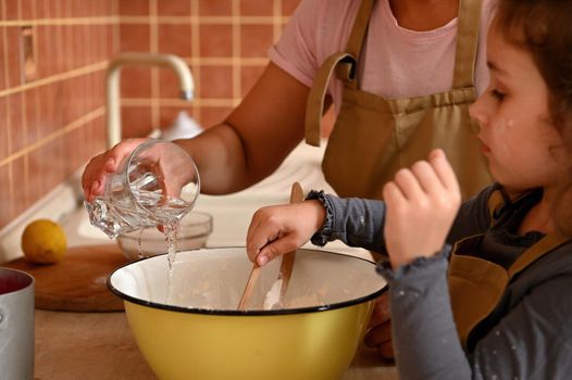 Selective focus on woman's hands, pouring water into vintage enamel bowl with white flour, standing near an adorable little girl kneading dough, while learning cooking delicious for festive cherry pie