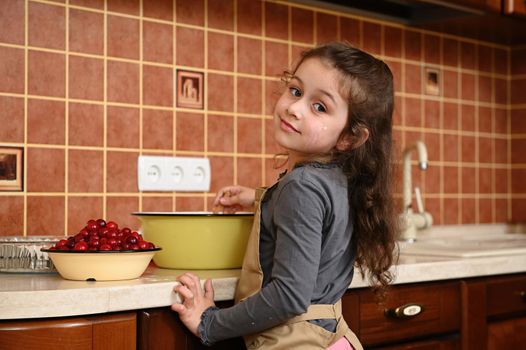 Portrait of an adorable Caucasian little girl with long hair, wearing a beige chef's apron, looking through her shoulder to the camera while kneading dough for preparing delicious homemeade cherry pie