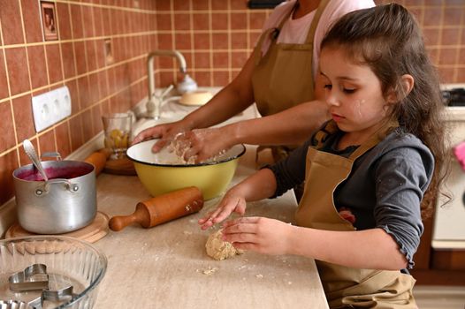 Beautiful mischievous Caucasian preschool child, cute baby girl, lovely daughter kneading dough on a kitchen countertop, learning cooking pastries and tartlets with her mom. Baking and cooking concept