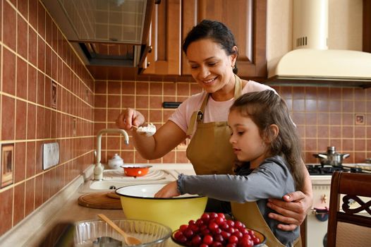 Beautiful African American young woman in chef apron, loving mom smiling and hugging her daughter, adorable multi-ethnic little child girl, while teaching her cooking homemade pie in the home kitchen