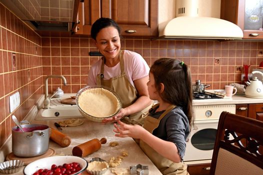 Cute little girl and her beautiful mom in chef's aprons, are playing and laughing while kneading the dough in the kitchen. Charming woman holding a glass mold with rolled dough for filling cherries
