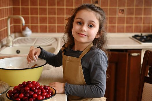 Pretty Caucasian child, blue-eyed baby girl wearing beige chef apron, standing by a coutertop with fresh ingredients, kneading dough, cooking a festive cherry cake, cutely smiling looking at camera