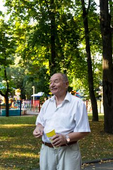 An elderly man walks alone in the park in the summer. A pensioner in a white shirt stands alone in the park with a glass of popcorn.