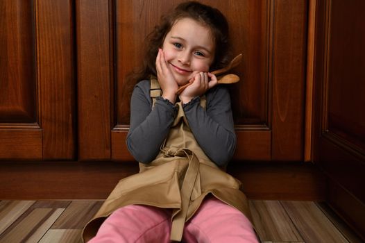 Adorable little girl in beige chef apron, sitting on the floor, leaning against wooden kitchen cupboard, holding cooking utensils, cutely smiling, looking to camera. Confectioner child, baker, cook