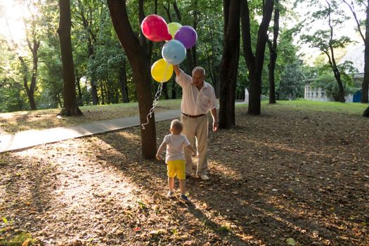 A little boy in yellow shorts and an elderly old man are playing with bright balloons. The family is having fun in the park in the fresh air and celebrating the holiday..