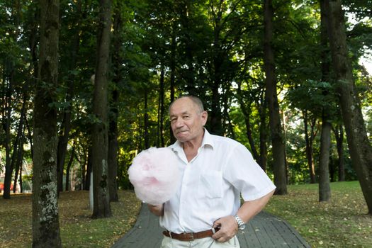 An old retired man in a white shirt walks with a huge, pink, cotton candy on the path in the park.