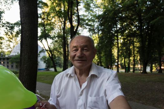 An elderly man with a smile on his face and bright balloons makes a selfie in the park.