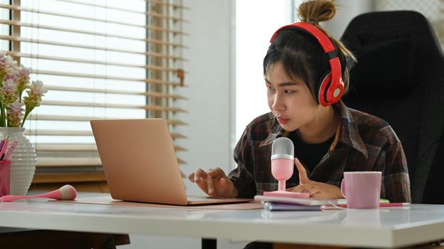 Young podcaster using a microphone and laptop computer recording audio for channel in home studio.