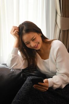 Satisfied woman resting on couch and using mobile phone phone, spending time in winter or autumn weekend at home.