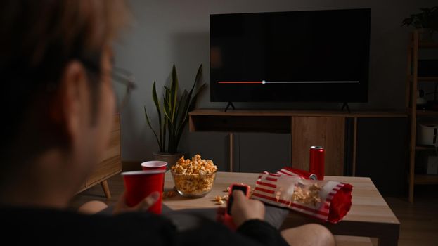 Happy man with remote control eating popcorn and watching movie in living room. Entertainment and leisure activity concept.
