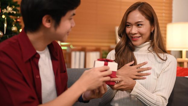Smiling man giving surprise gift to his girlfriend, celebrating Christmas at home. Family, holidays and people concept.