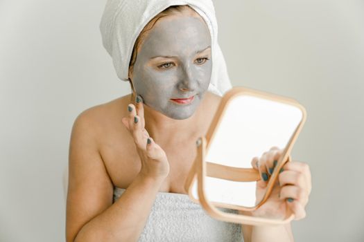 Girl wrapped in towel puts gray cosmetic clay mask on her face while looking in mirror. Close-up