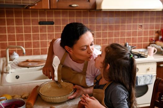 Beautiful dark-haired Hispanic young woman wearing beige chef apron, a loving mom kissing her cute baby, a lovely daughter while cooking together delicious tartlets and cherry pie in the home kitchen