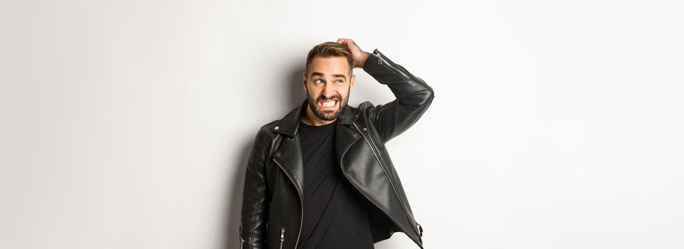 Confused macho guy in black leather jacket looking awkward and unsure, scratching head puzzled and stare left, white background.