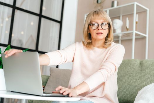 Thinking Freelance mature senior woman in glasses typing at laptop and working from home office with plants. Happy girl on workplace at the desk. Distance learning online education.