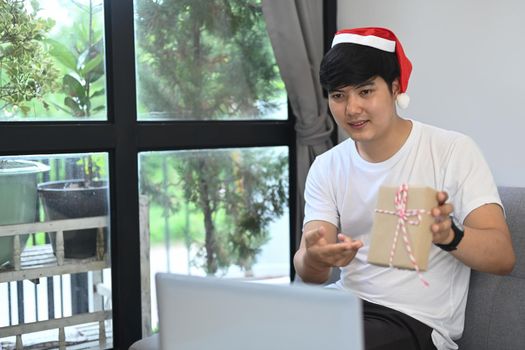 Happy young man in red Santa hat showing Christmas present during video call with his family at Christmas time.