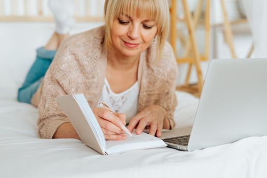 Distance learning online education and work. Mature adult woman lying on bed, writing in notebook. Freelancer with laptop at home office. Using computer and online shopping.