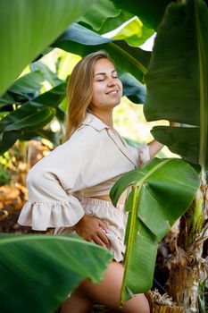 Woman near green leaves of banana bushes on nature in a park in a tropical place, she is in a beige skirt and a blouse