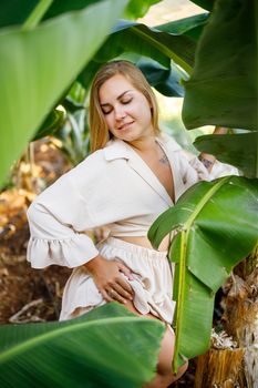 Woman near green leaves of banana bushes on nature in a park in a tropical place, she is in a beige skirt and a blouse