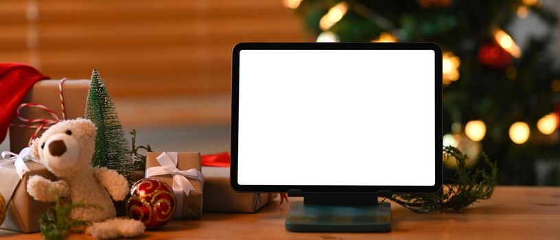 Digital tablet with blank screen and Christmas gift boxes on wooden table.