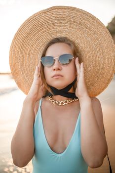 A young tanned woman in a beautiful swimsuit with a straw hat stands and rests on a tropical beach with sand and looks at the sunset and the sea. Selective focus. Vacation concept by the sea