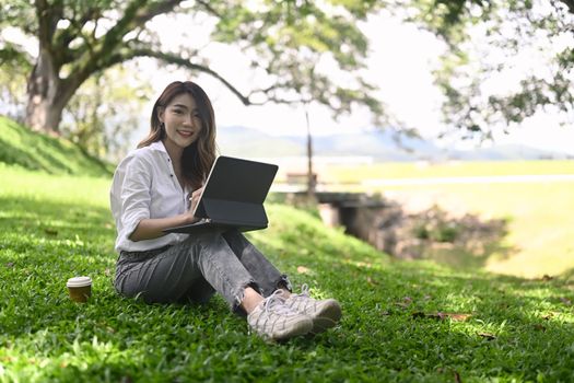 Happy woman sitting on grass with computer laptop in public park.