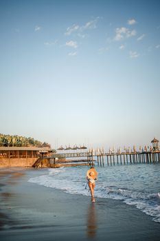 A woman in a swimsuit and a hat with glasses walks along the beach at sunset. The concept of sea recreation. Selective focus